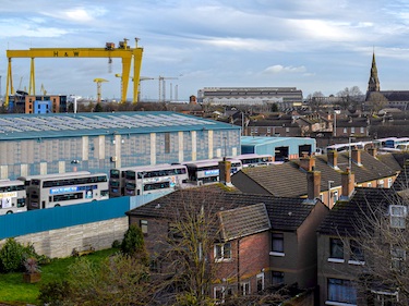 Skyline view of homes and industrial uses in Belfast, Northern Ireland. Two yellow shipbuilding gantry cranes are visible on the left behind a large warehouse and grey city buses. On the right, multiple church spires and red brick homes are shown, with garden trees in the foreground.
