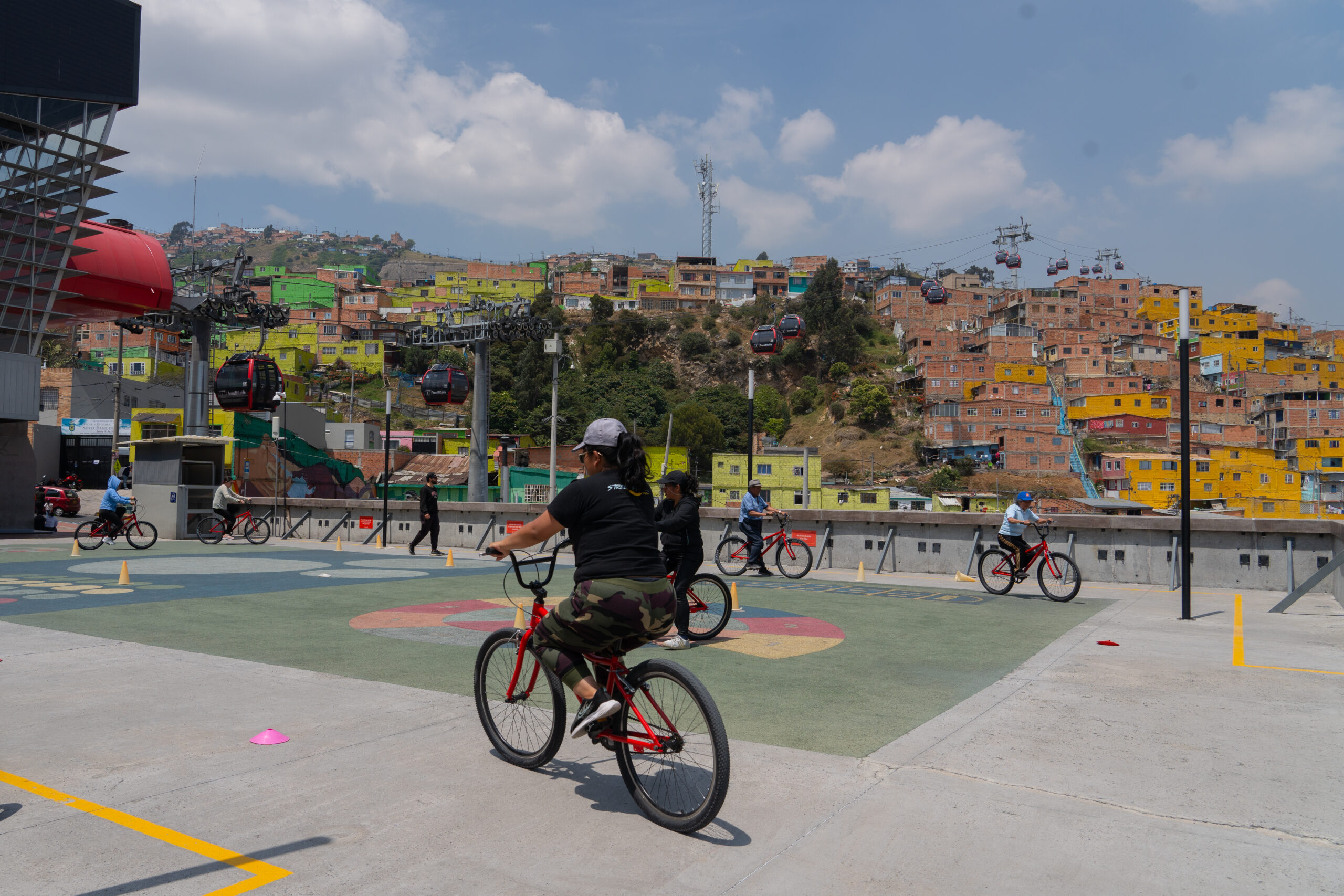 A group of people biking with a woman biking at the front of the picture. The platform that people bike on is surrounded by a hill with densely built houses. Cable cars are in the background.