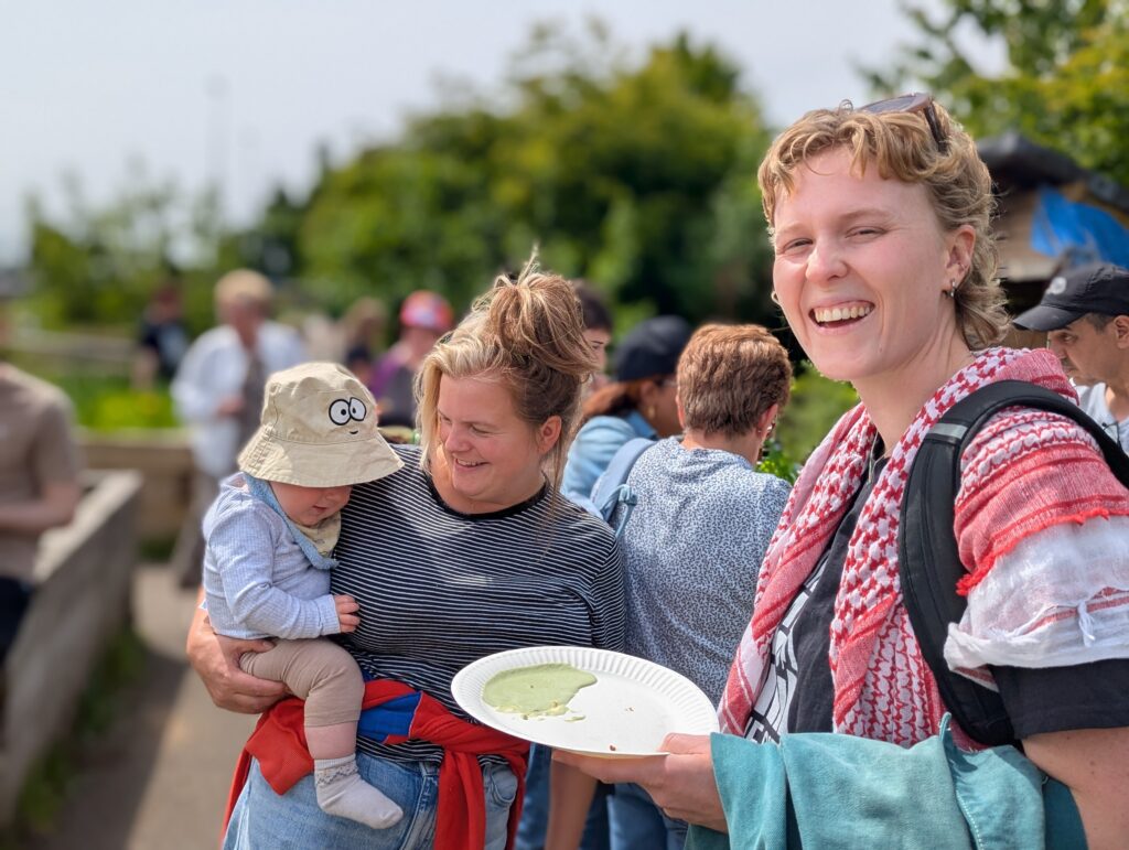 Two women and a child participating at an event in a Gairdín an Phobail (the People’s Garden). There are more people in the background and a lush natural environment.