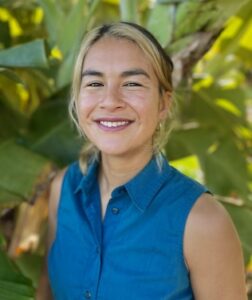 Photograph of Forest Abbott-Lum, self-described as "A photo of a smiling woman with light hair tied back, wearing a blue sleeveless button-down shirt standing in front of large green tropical leaves."