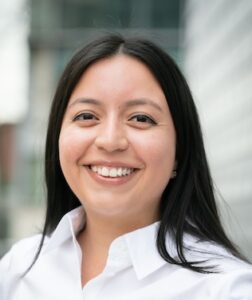 Photograph of Kelly Naranjo, self-described as "A portrait of a smiling woman with shoulder-length straight black hair wearing a white button-down shirt against a blurred architectural background."