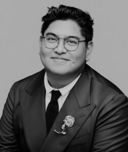 Photograph of Kevin Borja self-described as "A black and white portrait of a smiling man wearing round glasses and a dark suit with a tie and a rose lapel brooch."