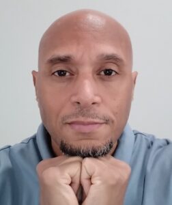 Photograph of Khalil Shahyd, self-described as "A portrait of a bald man with a goatee resting his chin on his clasped hands while looking directly at the camera, wearing a light blue polo shirt."