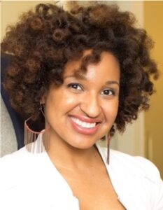 Vedette Gavin self-described her image as "A close-up of a smiling woman with fluffy curly brown hair and large dangling earrings, wearing a white top against a neutral background."